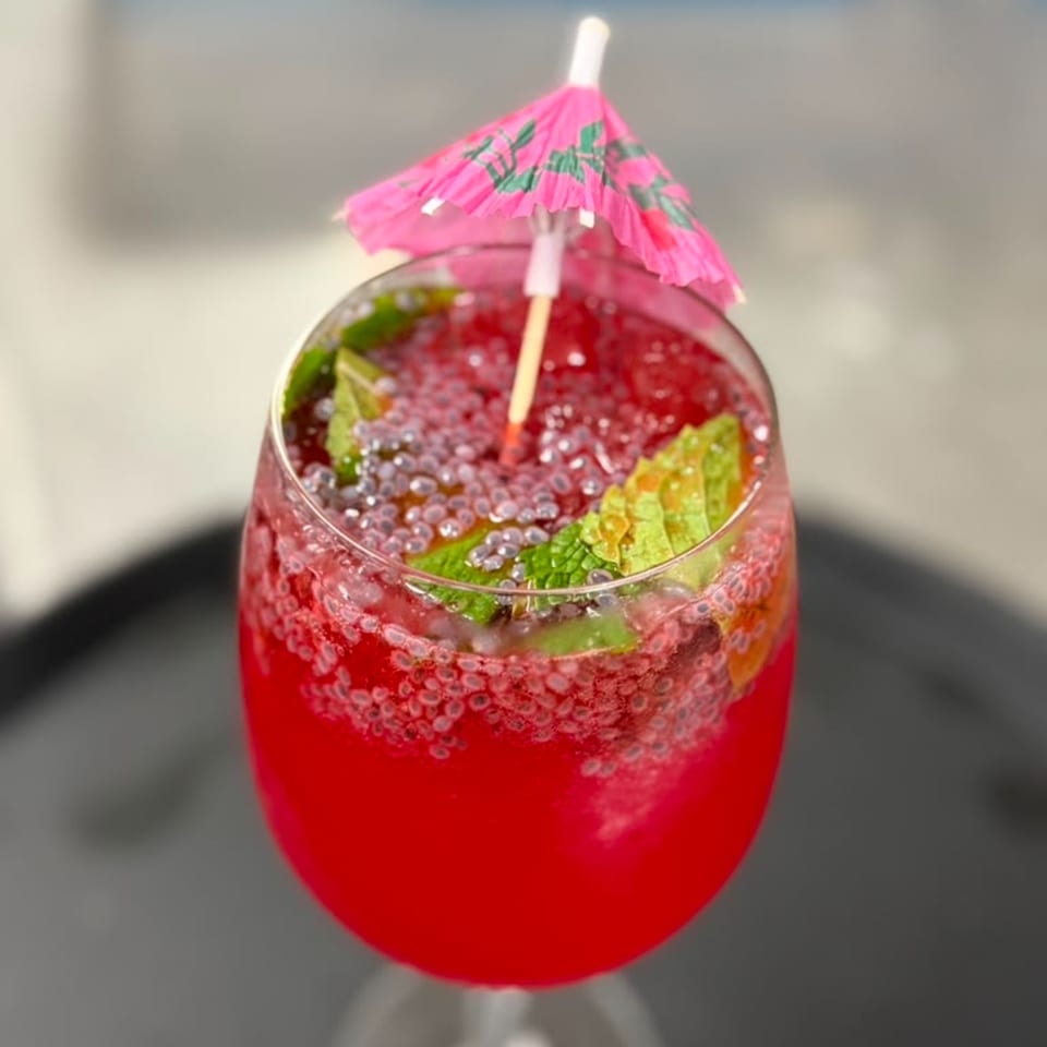 Bright red fruit cocktail with lime wedges, ice, bubbles, and a pink paper umbrella in a stemless glass.