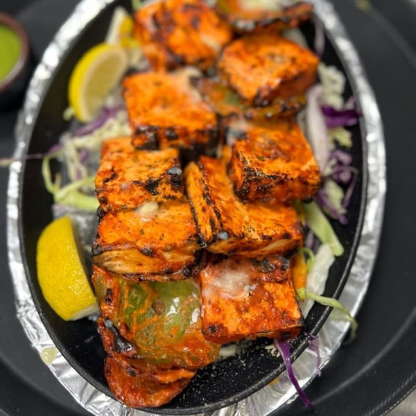 Sizzling Indian paneer tikka with bell peppers and lemon wedges on a black platter.