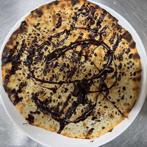 A round, golden-brown dessert pizza with dark chocolate drizzle and chocolate chips on a white plate.